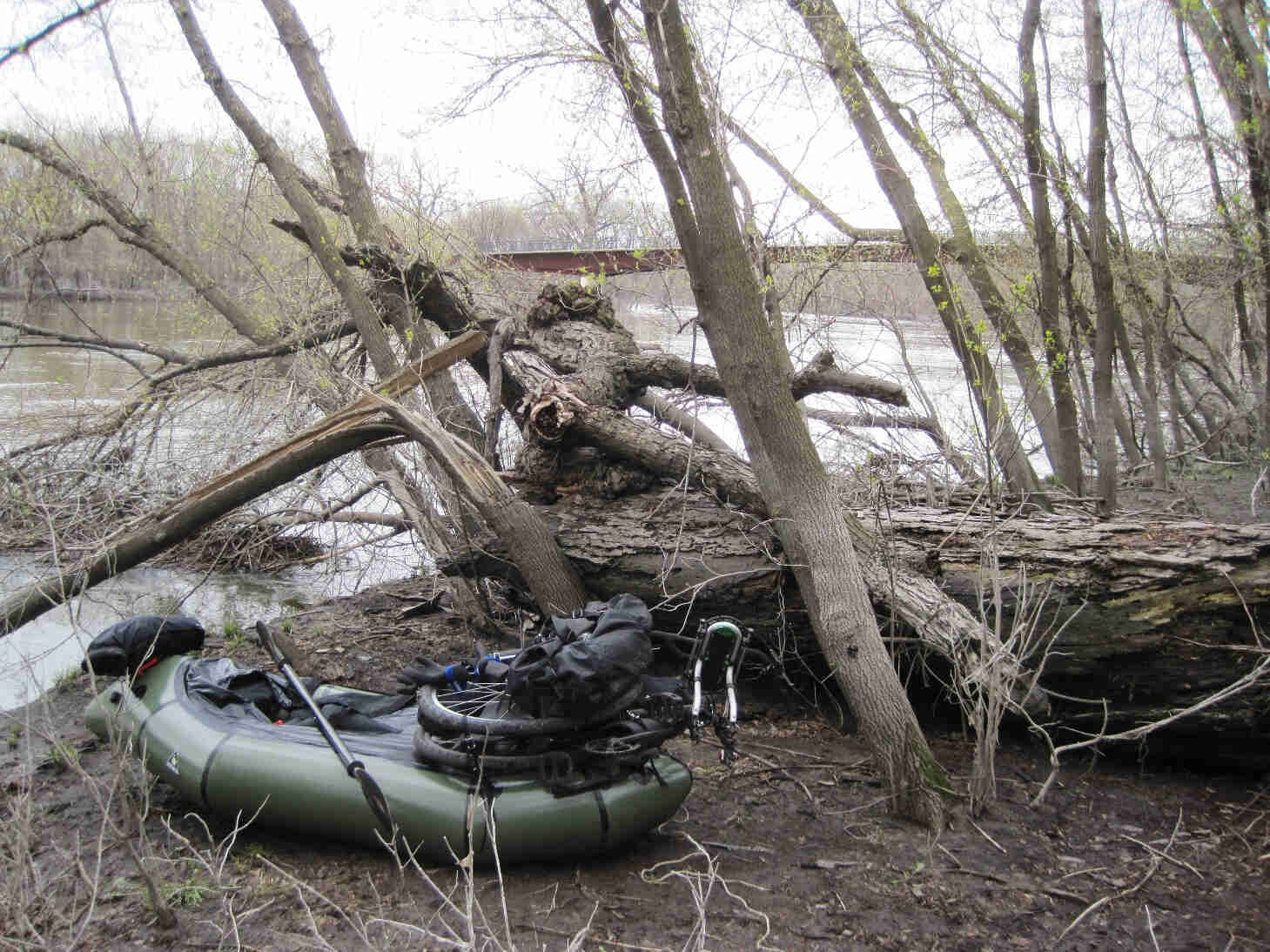 An olive green inflatable raft with a disassembled bike on top, on a dirt river bank in the bare woods